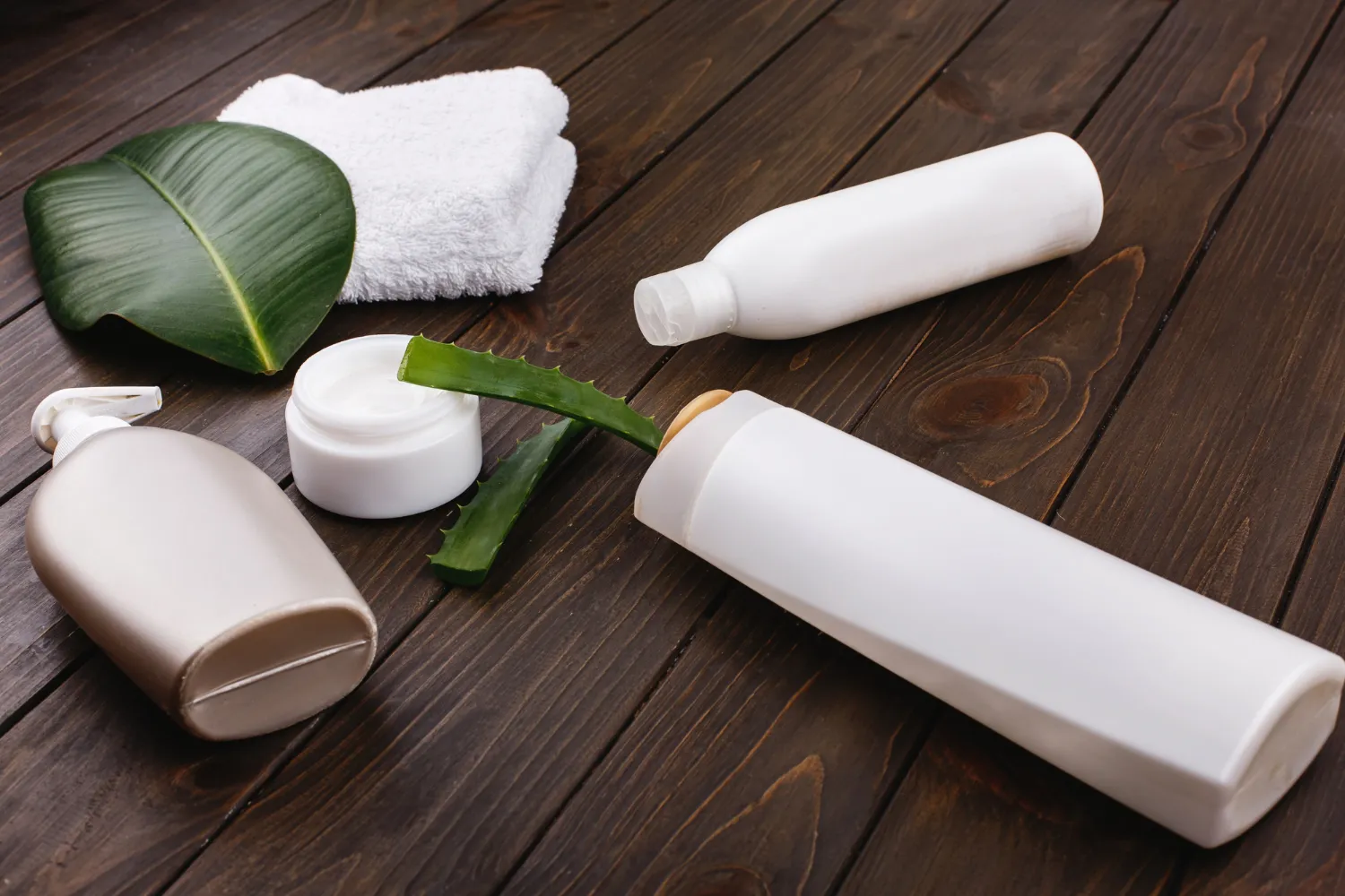 Productos de cuidado personal sobre una mesa de madera, incluyendo frascos de champú y acondicionador, una crema hidratante, una hoja de aloe vera y una toalla blanca, ideales para una rutina de belleza natural.